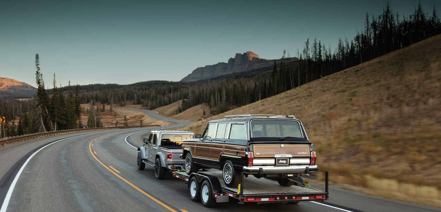 The 2021 Jeep Gladiator High Altitude towing a trailer with an older Jeep Grand Wagoneer on it.